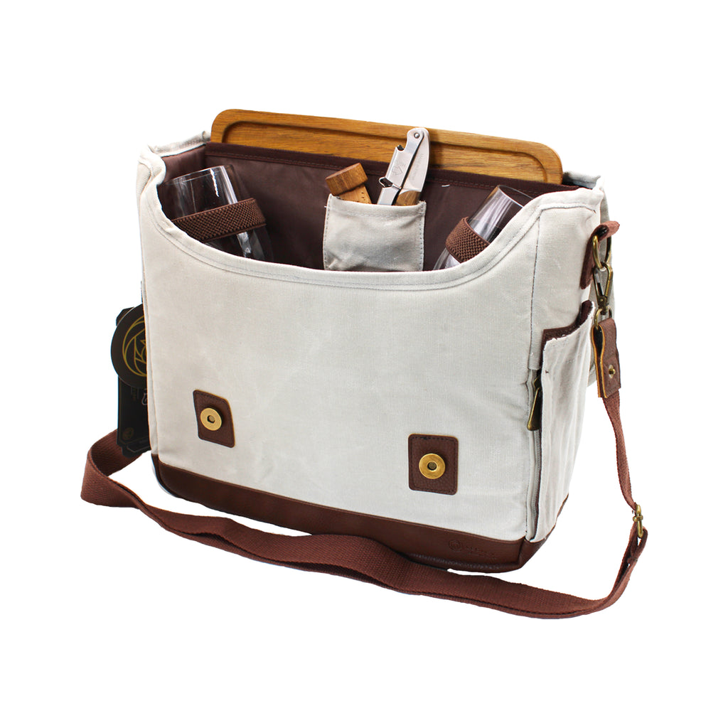 Image of an open off-white canvas tote bag with brown accents and wine bottle pocket on the side. Bag contains two wine glasses, a cork, a cork screw and a wooden cutting board.